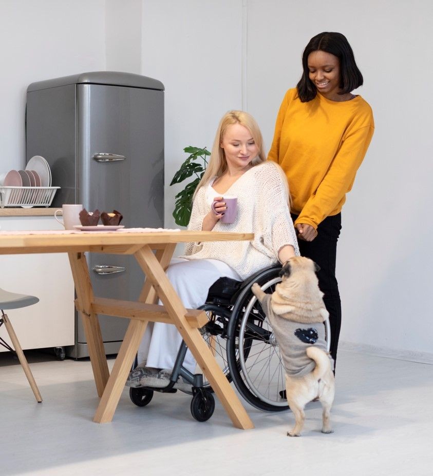 Woman in wheelchair and friend smiling at a small dog standing with paws on knee in kitchen.