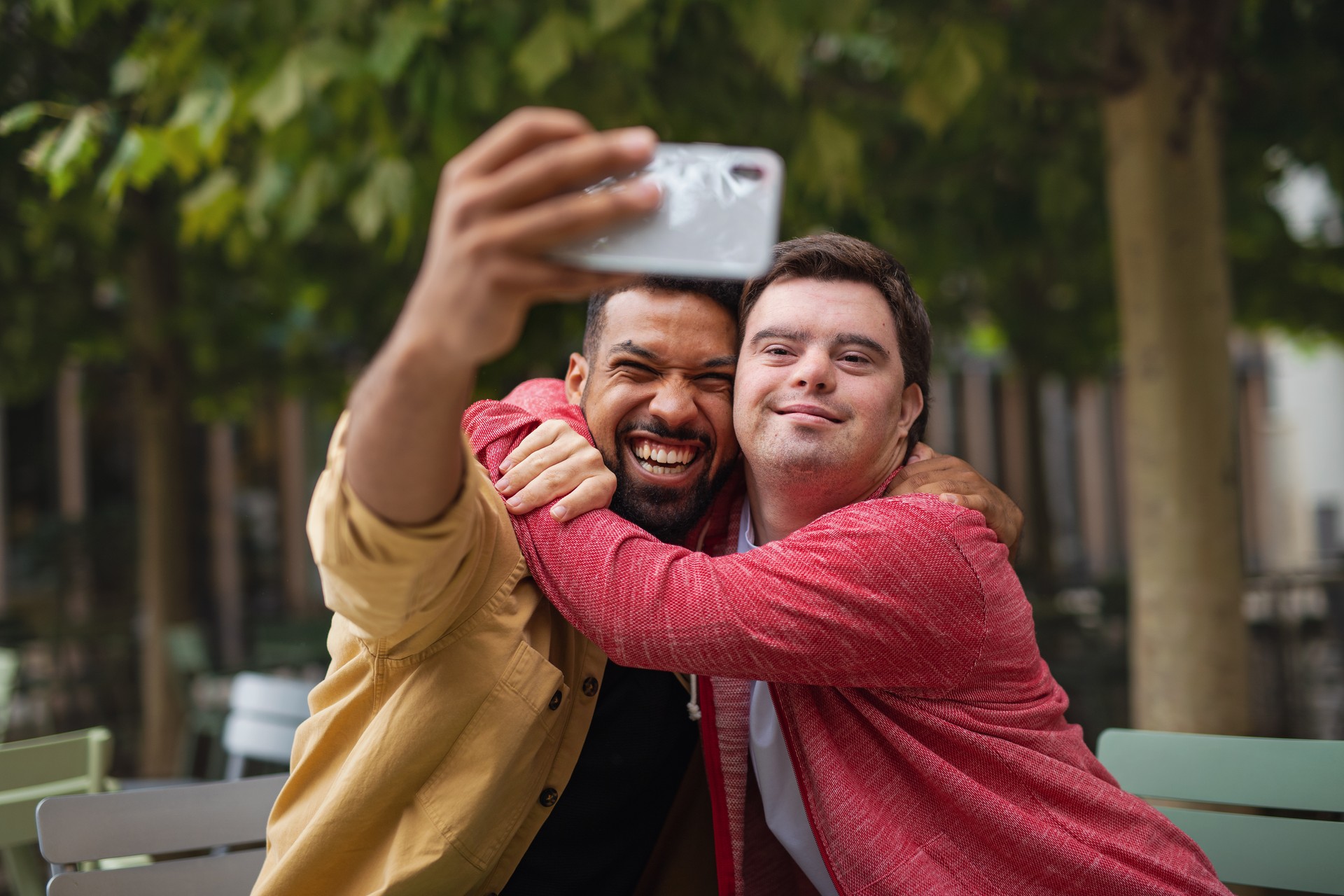 Young man with Down syndrome and his mentoring friend sitting and taking selfie outdoors in cafe